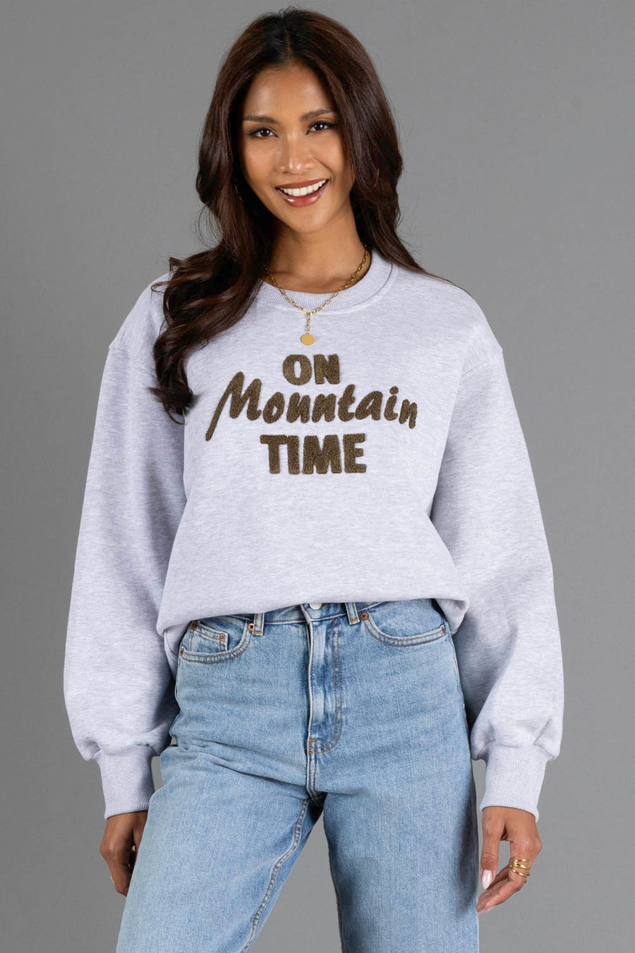 A woman with long brown hair smiles, wearing the Panache Apparel Collection Heather Gray & Brown On Mountain Time Sweatshirt and blue jeans, standing against a plain gray background.