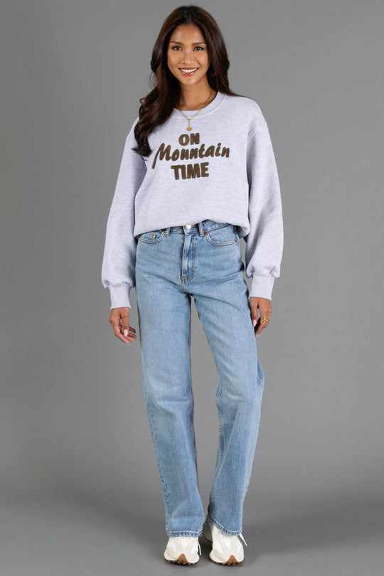 A woman stands and smiles, wearing the Panache Apparel Collection Heather Gray & Brown On Mountain Time Sweatshirt, paired with light blue jeans and white sneakers, set against a plain gray background.