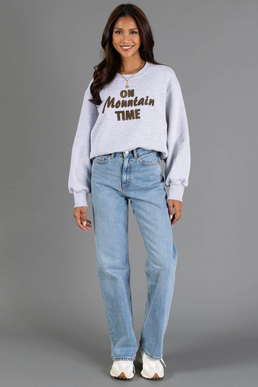 A woman stands and smiles, wearing the Panache Apparel Collection Heather Gray & Brown On Mountain Time Sweatshirt, paired with light blue jeans and white sneakers, set against a plain gray background.
