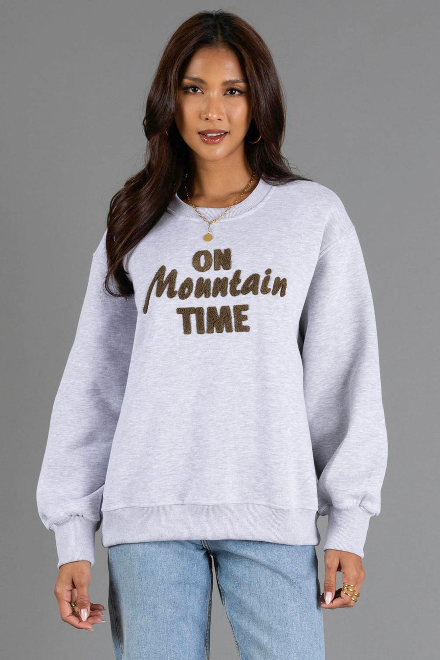 A woman with long dark hair is wearing the Panache Apparel Collection Heather Gray & Brown On Mountain Time Sweatshirt, paired with blue jeans, a gold necklace, and rings, standing against a plain gray background.