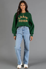A woman stands and smiles, dressed in the Panache Apparel Collection Green & Tan Cabin Fever Sweatshirt, paired with light blue jeans and white sneakers, set against a plain gray background.