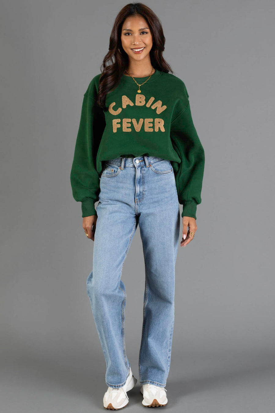 A woman stands and smiles, dressed in the Panache Apparel Collection Green & Tan Cabin Fever Sweatshirt, paired with light blue jeans and white sneakers, set against a plain gray background.