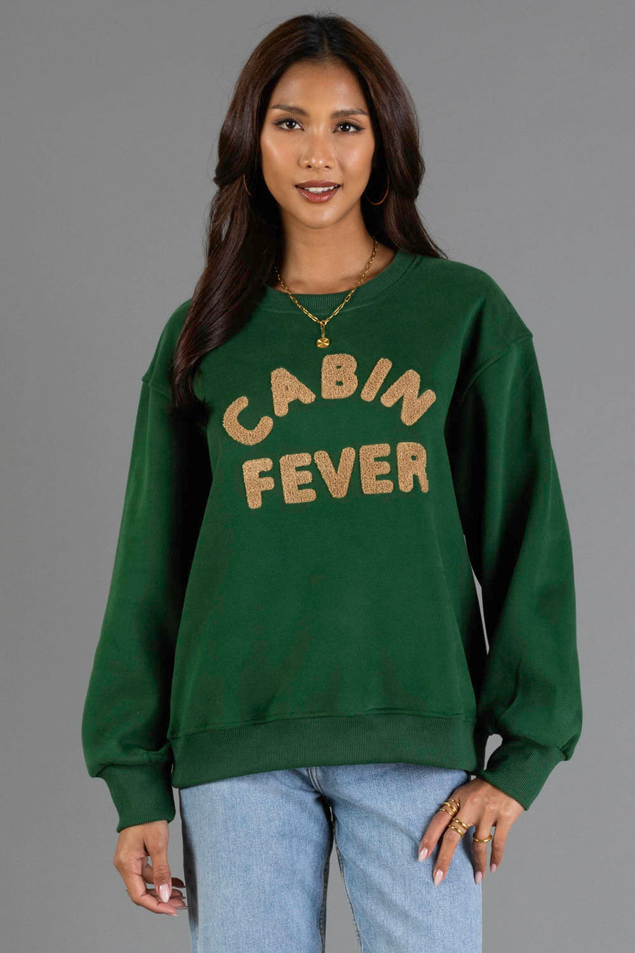 A woman stands facing the camera, modeling the Panache Apparel Collection’s Green & Tan Cabin Fever Sweatshirt. The sweatshirt features “CABIN FEVER” in tan letters on a green background. She has long brown hair, wears jeans, gold jewelry, and is posed against a plain gray backdrop.