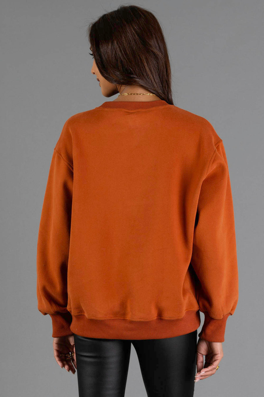 A person with long brown hair is seen from behind wearing Panache Apparel Collection’s Rust & Cream Cabin Fever Sweatshirt and black pants, standing against a plain gray background.