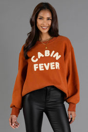 A woman is wearing the Panache Apparel Collection Rust & Cream Cabin Fever Sweatshirt, featuring oversized styling and cream chenille lettering, paired with black faux leather pants and a gold necklace.