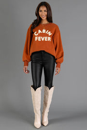 A woman stands and smiles against a plain gray background, wearing the Panache Apparel Collection Rust & Cream Cabin Fever Sweatshirt, paired with black shiny pants and tall cream cowboy boots.