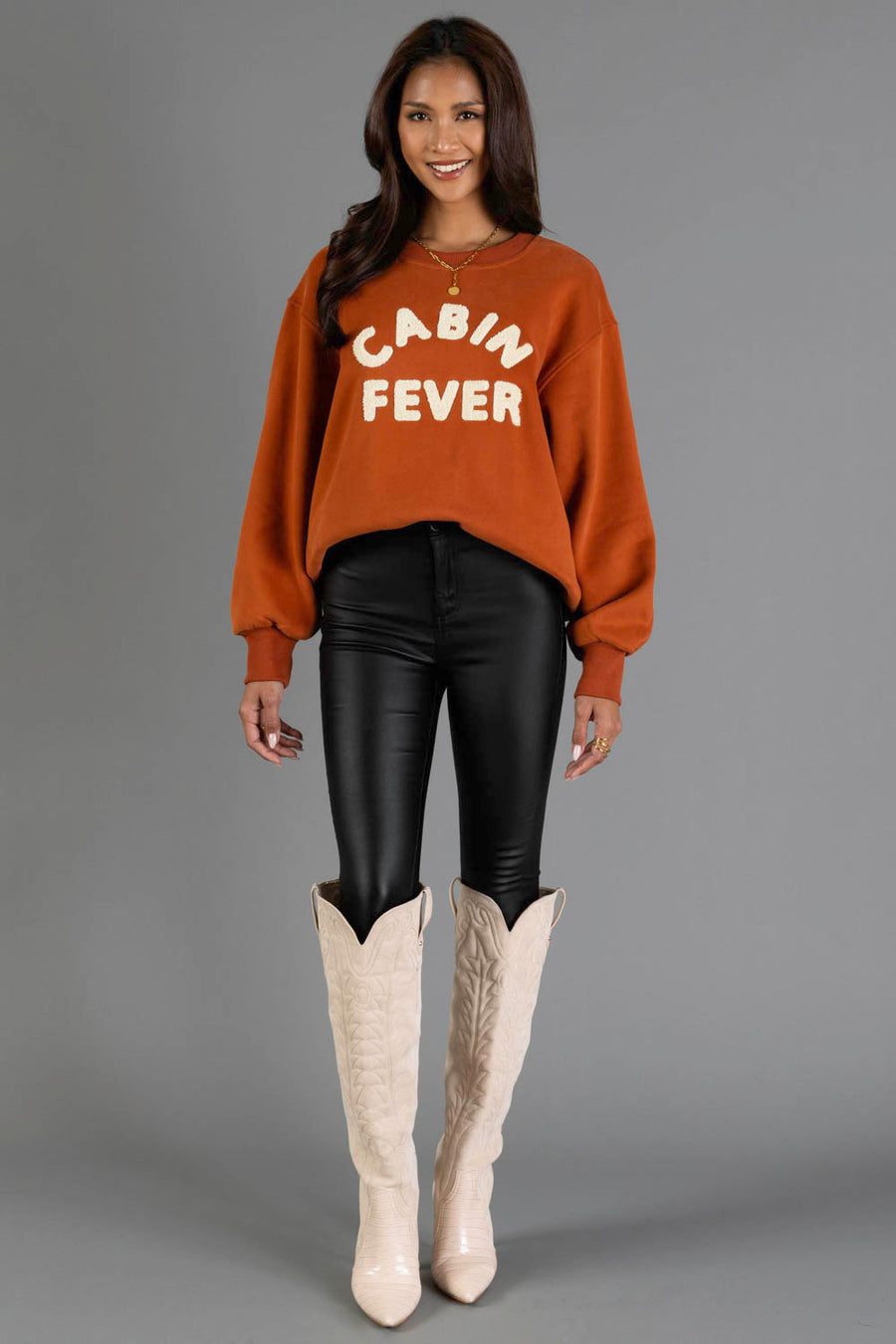 A woman stands and smiles against a plain gray background, wearing the Panache Apparel Collection Rust & Cream Cabin Fever Sweatshirt, paired with black shiny pants and tall cream cowboy boots.
