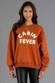 A woman with long dark hair wears the Panache Apparel Collection’s Rust & Cream Cabin Fever Sweatshirt, featuring “CABIN FEVER” in white letters, paired with black leather pants. She smiles as she stands against a plain gray background.