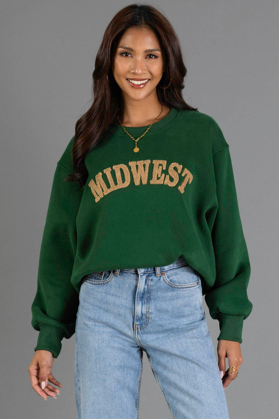 A woman with long dark hair is smiling and standing against a plain gray background, wearing the Panache Apparel Collection’s Green & Tan Midwest Sweatshirt paired with light blue jeans.