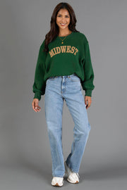 A woman stands and smiles against a plain gray background, wearing the Panache Apparel Collection Green & Tan Midwest Sweatshirt, paired with light blue jeans and white sneakers.