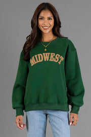 A woman with long dark hair wears the Green & Tan Midwest Sweatshirt from Panache Apparel Collection and light blue jeans, standing and smiling against a plain gray background.