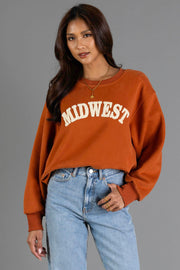 A person wearing the Panache Apparel Collection’s Rust & Cream Midwest Sweatshirt with light blue jeans stands against a gray background, one hand in their pocket and the other relaxed by their side.