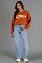 A woman wearing a Rust & Cream Midwest Sweatshirt from Panache Apparel Collection, paired with light blue jeans and white shoes, stands against a plain gray background, looking at the camera with a relaxed expression.