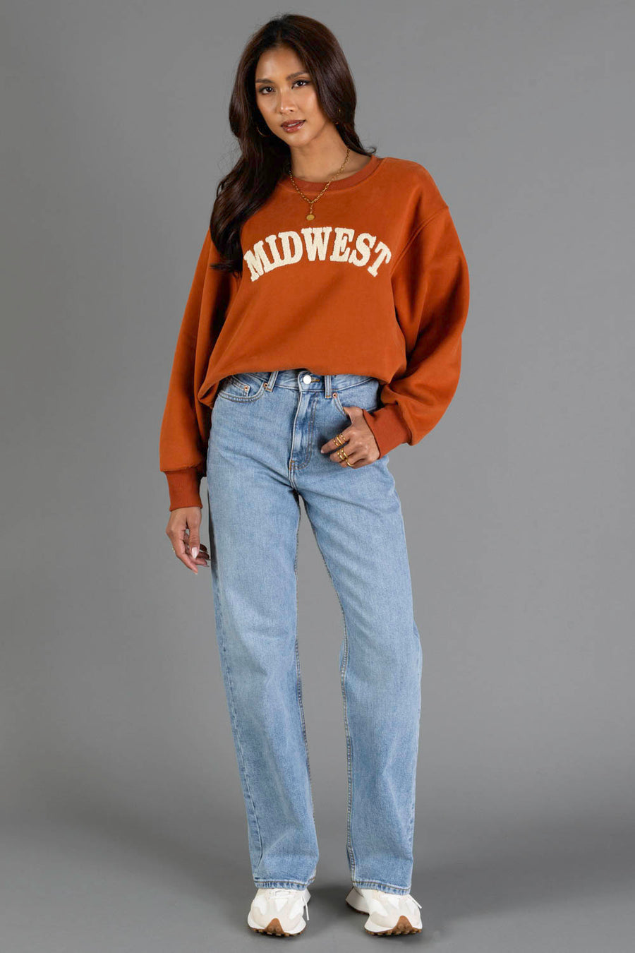 A woman wearing a Rust & Cream Midwest Sweatshirt from Panache Apparel Collection, paired with light blue jeans and white shoes, stands against a plain gray background, looking at the camera with a relaxed expression.
