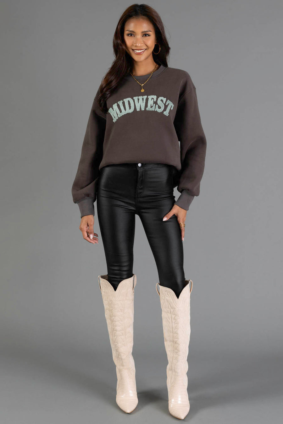 A woman stands and smiles, wearing the Charcoal & Mint Midwest Sweatshirt from Panache Apparel Collection, paired with black leather pants and tall white cowboy boots. Her hands rest relaxed by her sides against a plain gray background.