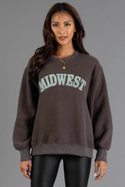A woman with long dark hair wears hoop earrings, a gold necklace, and the Panache Apparel Collection’s Charcoal & Mint Midwest Sweatshirt—an oversized dark sweatshirt featuring “MIDWEST” in large light letters—paired with black leather pants, standing against a plain gray background.