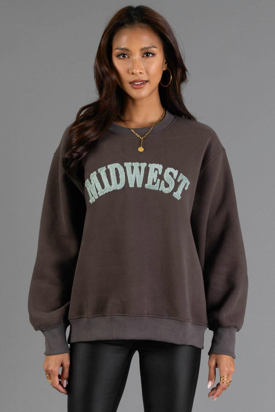 A woman with long dark hair wears hoop earrings, a gold necklace, and the Panache Apparel Collection’s Charcoal & Mint Midwest Sweatshirt—an oversized dark sweatshirt featuring “MIDWEST” in large light letters—paired with black leather pants, standing against a plain gray background.