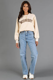 A woman wearing a Panache Apparel Collection Tan & Dark Brown Midwest Sweatshirt with balloon sleeves, light wash straight-leg jeans, and white sneakers stands smiling against a gray background.