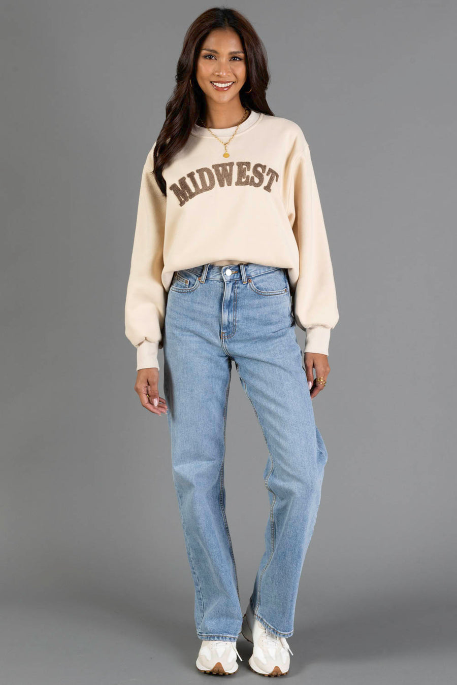 A woman wearing a Panache Apparel Collection Tan & Dark Brown Midwest Sweatshirt with balloon sleeves, light wash straight-leg jeans, and white sneakers stands smiling against a gray background.