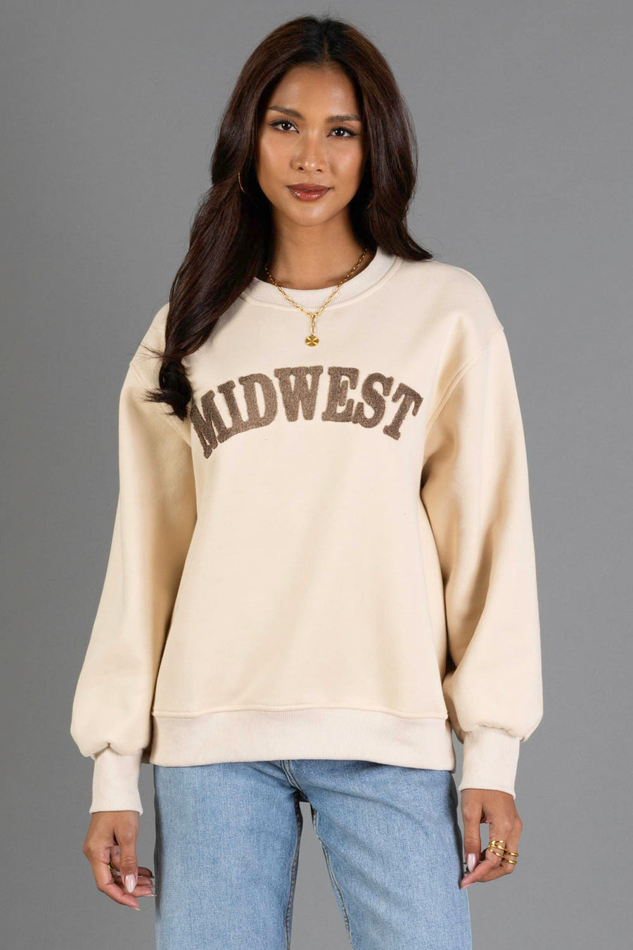 A woman with long dark hair wears a Panache Apparel Collection Tan & Dark Brown Midwest Sweatshirt, blue jeans, and gold jewelry as she stands against a gray background, smiling softly at the camera.