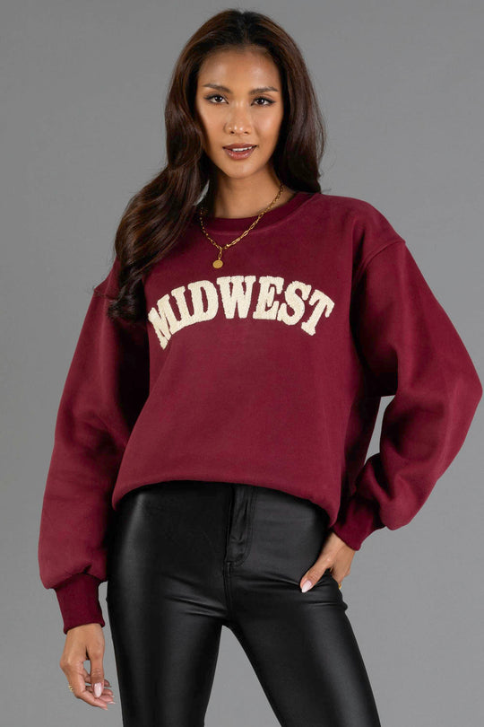 A woman with long brown hair wears the Panache Apparel Collection’s Maroon & Cream Midwest Sweatshirt paired with black leather pants, standing against a plain gray background.