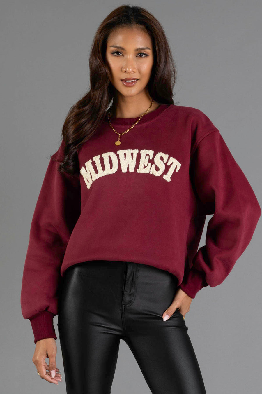 A woman with long brown hair wears the Panache Apparel Collection’s Maroon & Cream Midwest Sweatshirt paired with black leather pants, standing against a plain gray background.