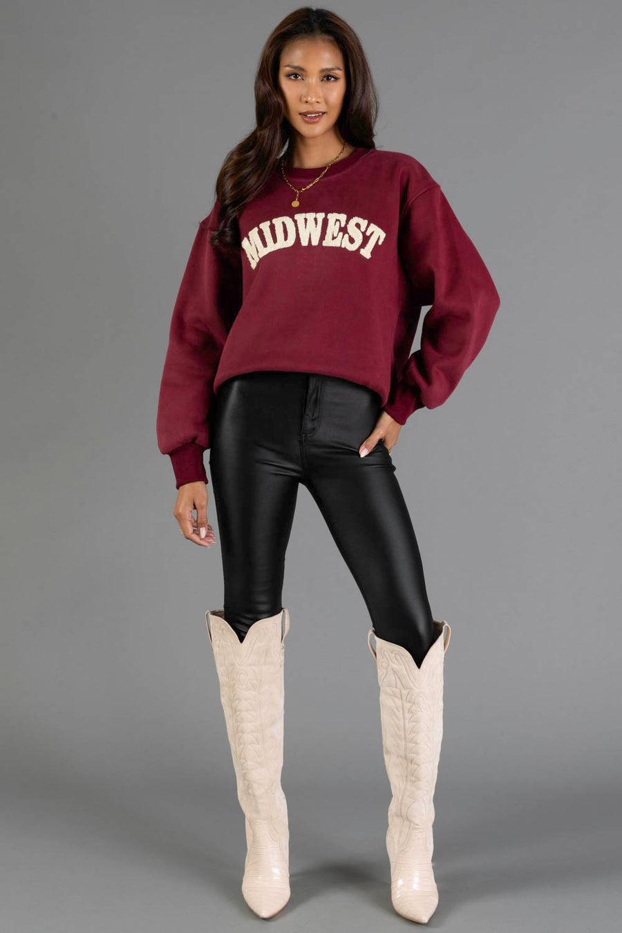 A woman poses against a plain gray background wearing the Panache Apparel Collection’s Maroon & Cream Midwest Sweatshirt, paired with black leather pants and white cowboy boots.