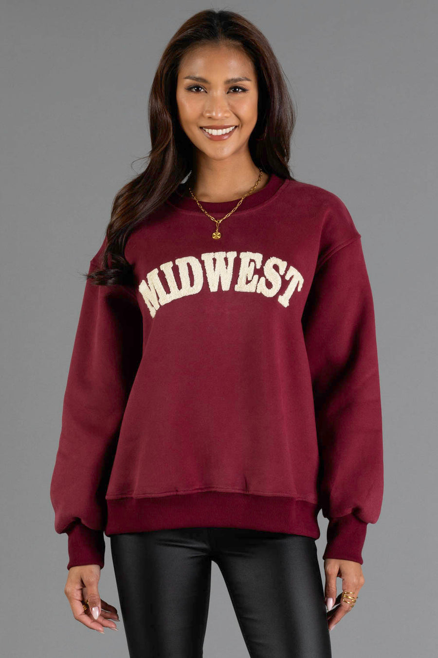 A woman wearing the Maroon & Cream Midwest Sweatshirt from Panache Apparel Collection, featuring “MIDWEST” printed in white letters, stands against a gray background, smiling with her hands by her sides.