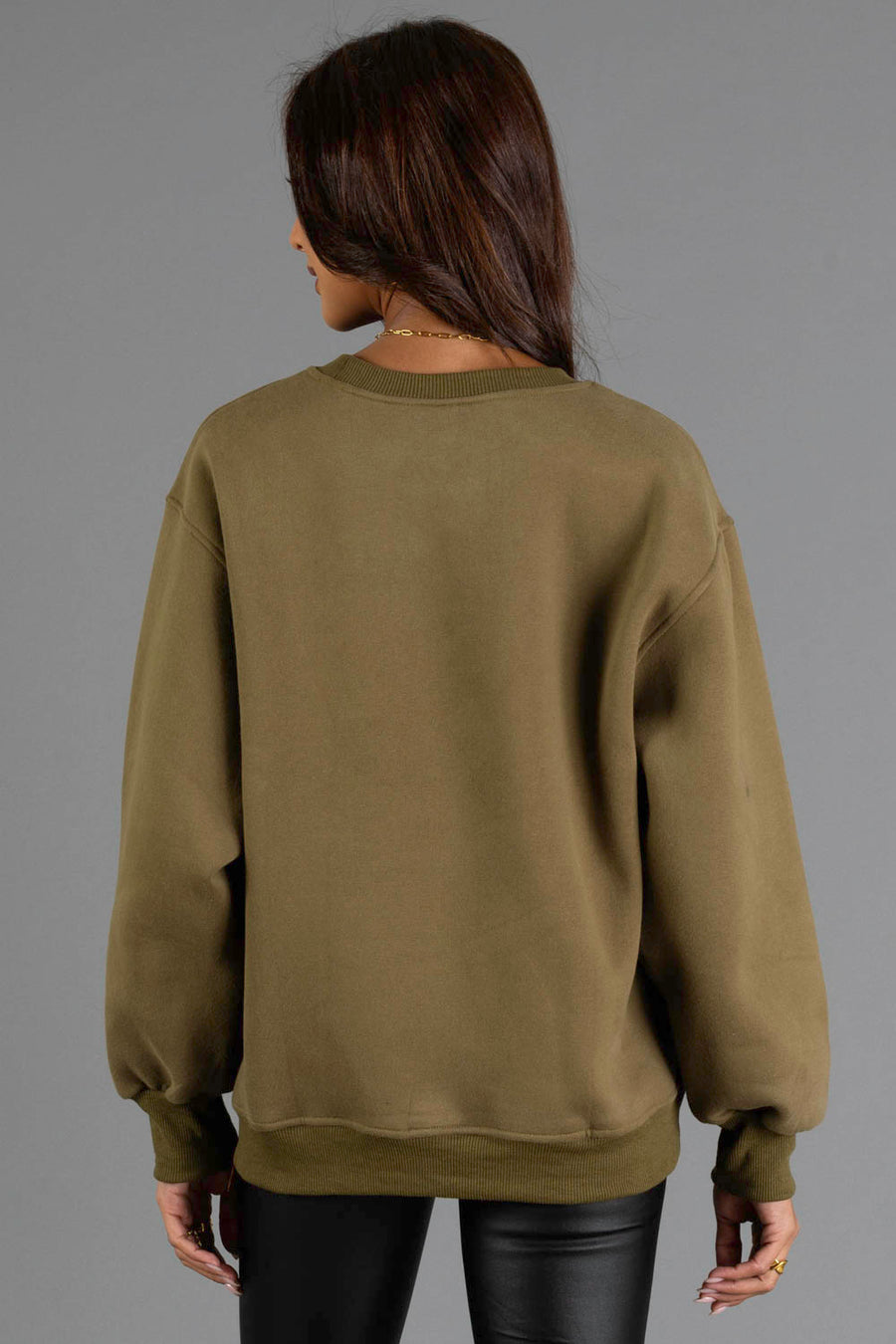 A woman with long brown hair wears the Olive & Beige Midwest Sweatshirt from Panache Apparel Collection and black leather pants, facing away from the camera against a plain gray background.