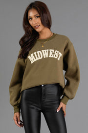 A woman wearing the Olive & Beige Midwest Sweatshirt from Panache Apparel Collection and black pants stands against a plain gray background. She has long hair, wears layered necklaces, and is smiling slightly.