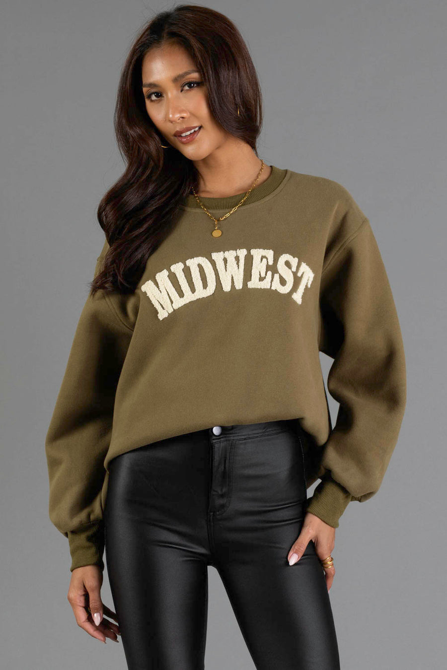 A woman wearing the Olive & Beige Midwest Sweatshirt from Panache Apparel Collection and black pants stands against a plain gray background. She has long hair, wears layered necklaces, and is smiling slightly.