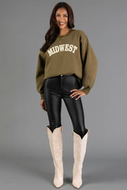 A woman stands smiling against a plain gray background, wearing a Panache Apparel Collection Olive & Beige Midwest Sweatshirt, black leather-look leggings, and tall white cowboy boots.