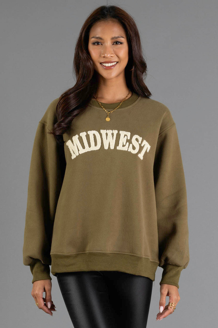 A woman with long brown hair smiles while wearing the Panache Apparel Collection’s Olive & Beige Midwest Sweatshirt, featuring “MIDWEST” in large white letters, paired with black leather pants and standing against a plain gray background.