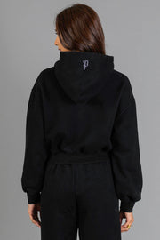 A person with long brown hair, dressed in the Black Panache Cropped Hooded Sweatshirt from the Panache Apparel Collection and matching black pants, faces away from the camera. A small white Panache logo is visible on the back of the hood. The plain gray background provides a simple backdrop.