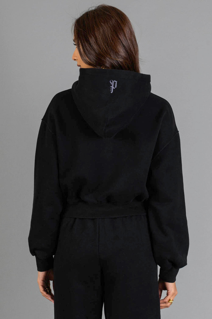 A person with long brown hair, dressed in the Black Panache Cropped Hooded Sweatshirt from the Panache Apparel Collection and matching black pants, faces away from the camera. A small white Panache logo is visible on the back of the hood. The plain gray background provides a simple backdrop.