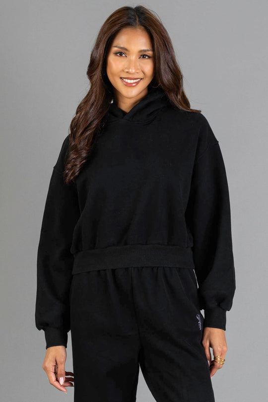 A woman with long brown hair is seen smiling against a plain gray background, wearing the Black Panache Cropped Hooded Sweatshirt and matching pants from Panache Apparel Collection.