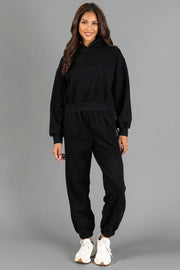 A person stands smiling, wearing the Black Panache Cropped Hooded Sweatshirt from the Panache Apparel Collection and matching jogger sweatpants with white sneakers, against a plain gray background.