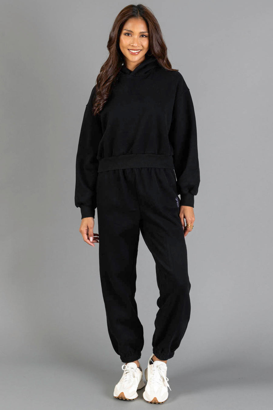 A person stands smiling, wearing the Black Panache Cropped Hooded Sweatshirt from the Panache Apparel Collection and matching jogger sweatpants with white sneakers, against a plain gray background.