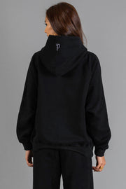 A person with long brown hair is seen from behind against a gray background, wearing the Panache Apparel Collection’s Black Panache Hooded Sweatshirt and black pants.