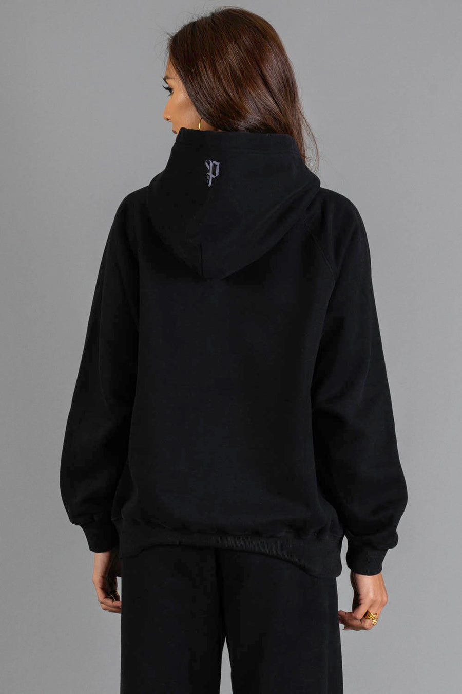 A person with long brown hair is seen from behind against a gray background, wearing the Panache Apparel Collection’s Black Panache Hooded Sweatshirt and black pants.