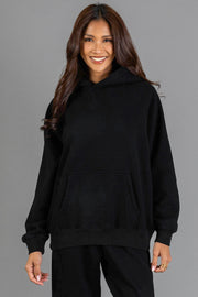 A person with long brown hair smiles while wearing the Black Panache Hooded Sweatshirt from the Panache Apparel Collection and black pants, standing against a plain gray background.