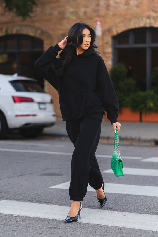 A person wearing a Black Panache Hooded Sweatshirt and matching black joggers walks across a crosswalk while carrying a bright green quilted handbag.