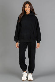 A woman stands smiling against a plain gray background, wearing the Black Panache Hooded Sweatshirt from Panache Apparel Collection, paired with black jogger pants and white sneakers.