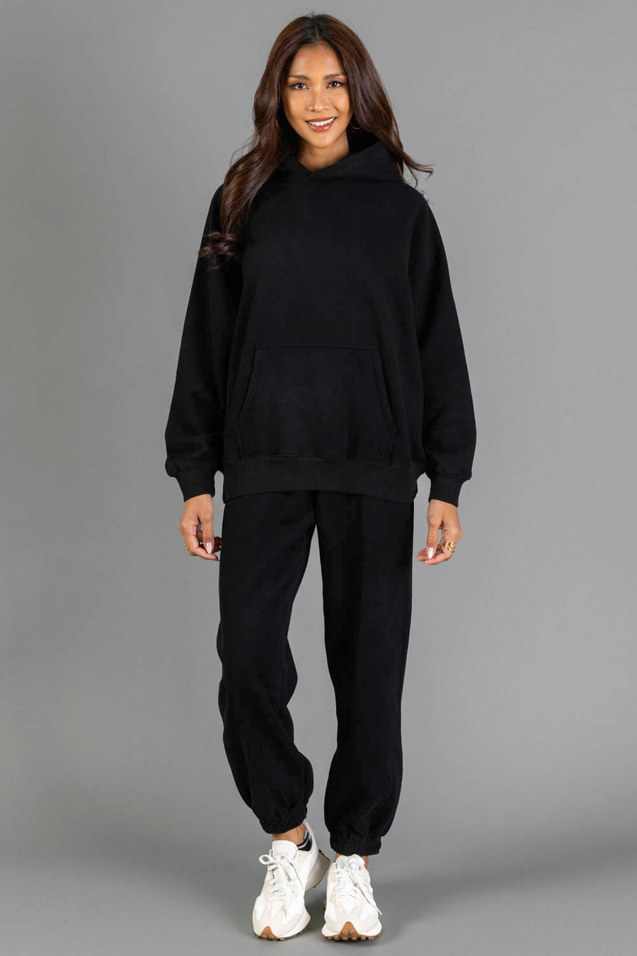 A woman stands smiling against a plain gray background, wearing the Black Panache Hooded Sweatshirt from Panache Apparel Collection, paired with black jogger pants and white sneakers.