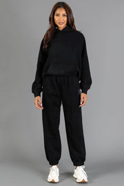A woman stands against a gray background wearing the Black Panache Hooded Sweatshirt from the Panache Apparel Collection, paired with matching black jogger pants and white sneakers. She is smiling slightly, and her long, wavy hair frames her face.