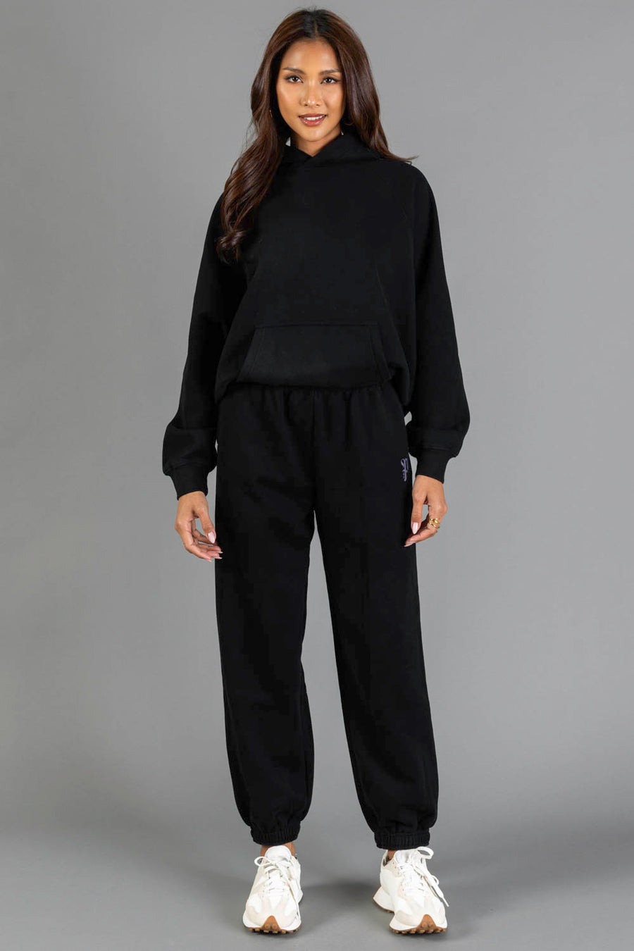 A woman stands against a gray background wearing the Black Panache Hooded Sweatshirt from the Panache Apparel Collection, paired with matching black jogger pants and white sneakers. She is smiling slightly, and her long, wavy hair frames her face.