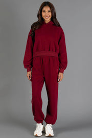 A woman stands smiling against a plain gray background, wearing the Maroon Panache Sweatpants from the Panache Apparel Collection styled with a matching hoodie and white sneakers.
