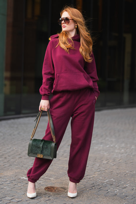 A woman wears a Maroon Panache Hooded Sweatshirt with matching maroon sweatpants, white heels, and a dark green handbag.