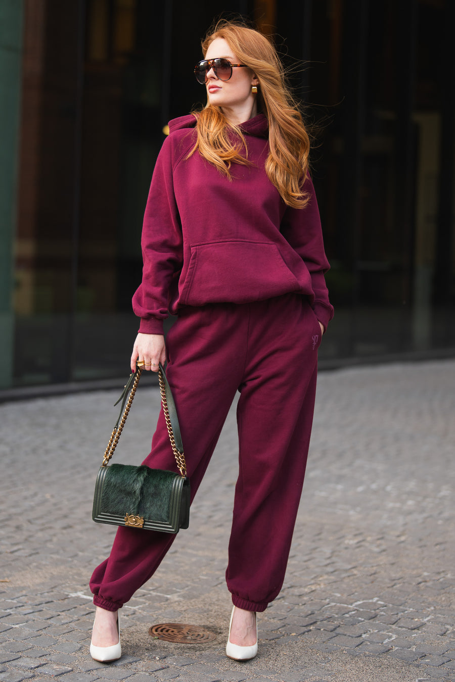A woman wears a Maroon Panache Hooded Sweatshirt with matching maroon sweatpants, white heels, and a dark green handbag.