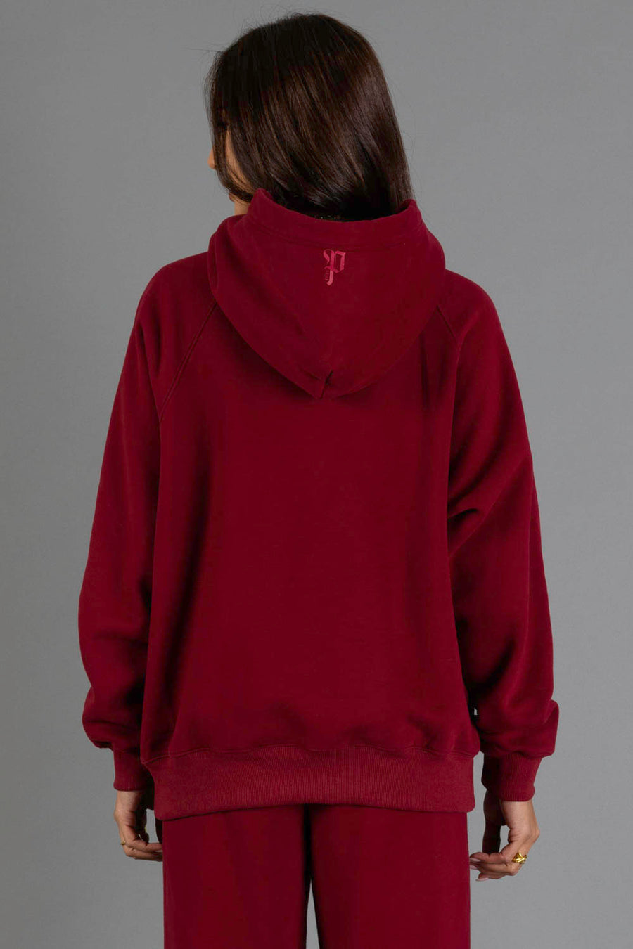 A person stands facing away, wearing the Maroon Panache Hooded Sweatshirt and matching pants from the Panache Apparel Collection against a plain gray background.